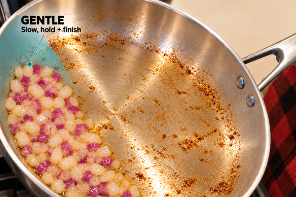 Close-up of a stainless steel sloped skillet on a stovetop, showing a portion of diced onions and garlic cooking while the rest of the pan has browned residue. Text in the upper left reads 'GENTLE Slow, hold + finish'.